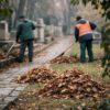 Vue de dos, des ouvriers ratissent les feuilles pour les mettre en tas près des chemins de pierre pour le nettoyage des débris à l'extérieur.