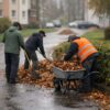 Contractor-style leaf cleanup scaling: workers raking and wheelbarrow loading in European grounds maintenance, autumn.