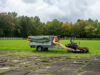 Zero-turn mower connected by suction hose to a Foresteel FT-COMBIvac vacuum trailer on a football pitch.
