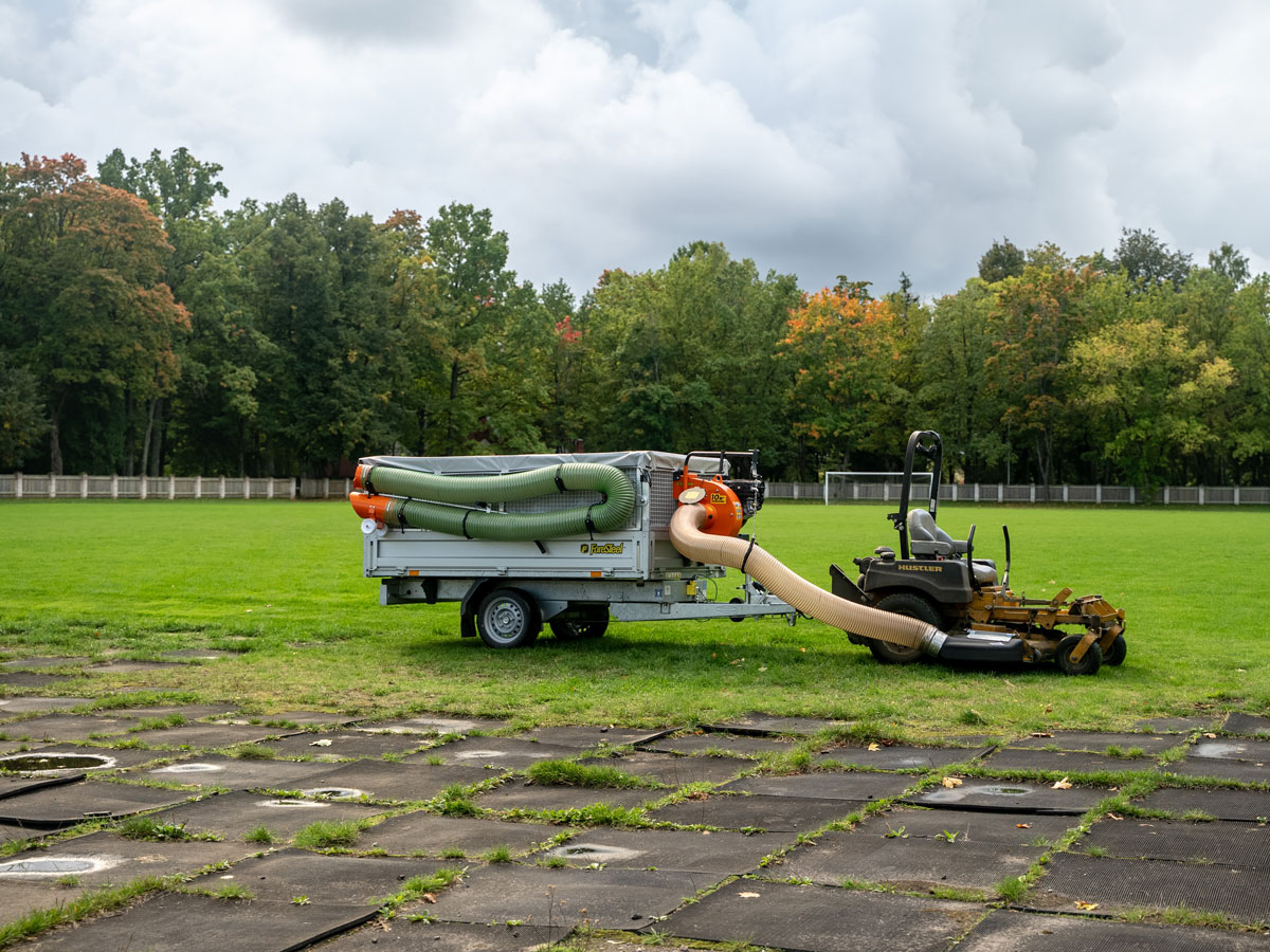 Zero-turn mower connected by suction hose to a Foresteel FT-COMBIvac vacuum trailer on a football pitch.
