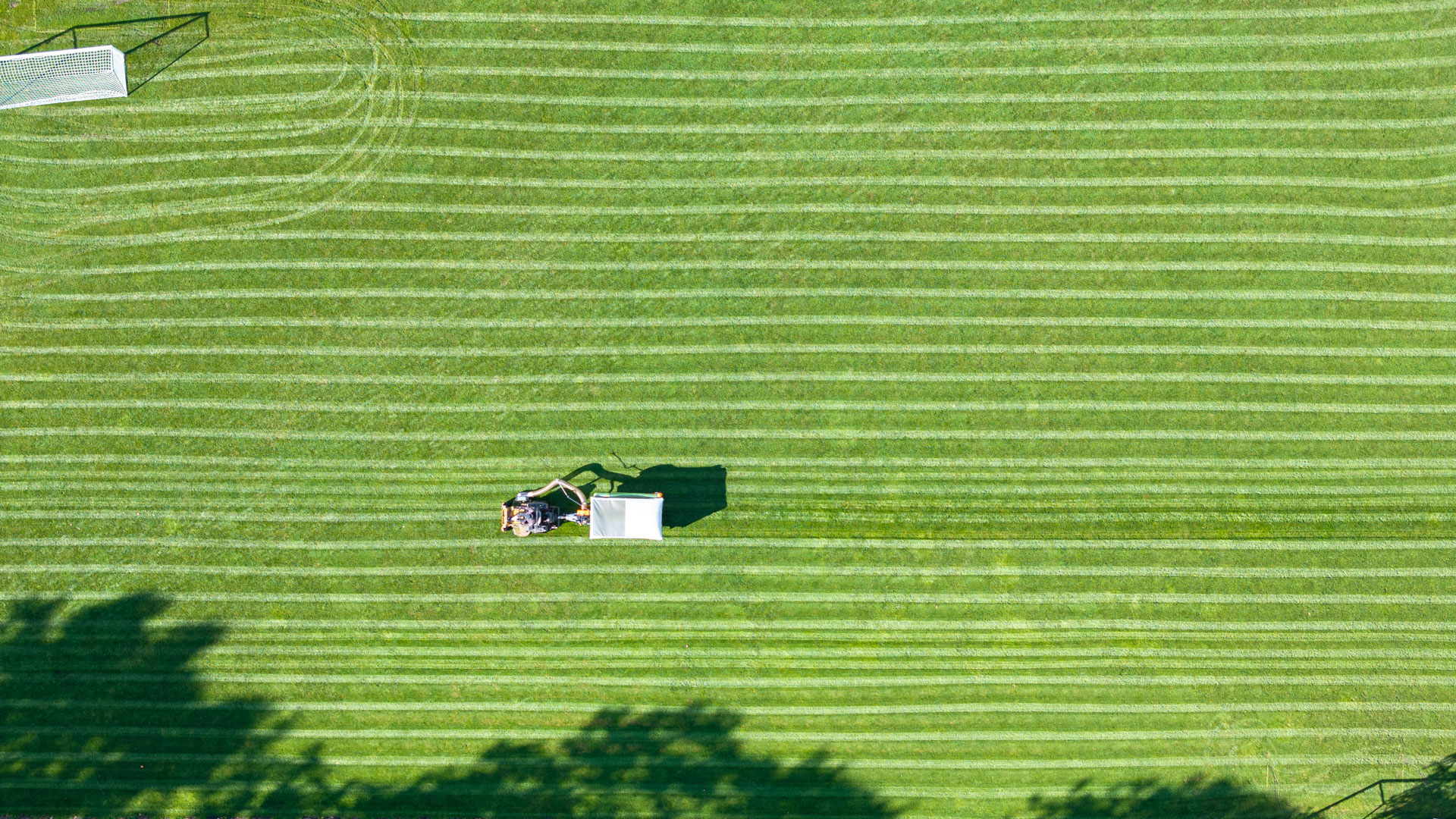 Aerial view of a football pitch being mowed and collected in one pass, showing cut and uncut sections.